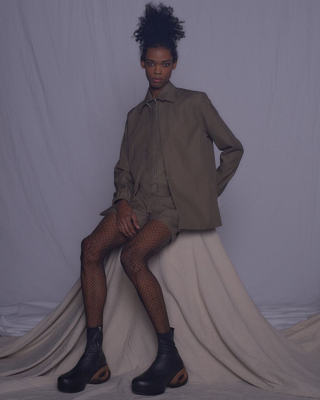 Woman poses in a studio wearing a beige zip-up jacket, matching shorts, fishnet tights, and chunky black boots against a neutral backdrop.