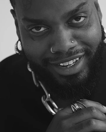 Close-up monochrome portrait of a Black man with facial tattoos, nose rings, and grillz, photographed by Katsu Naito.