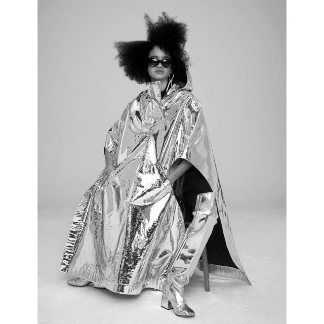 A person sits in a chair wearing a silver, hooded, metallic raincoat and matching, shiny knee-high boots in a black and white studio shot.