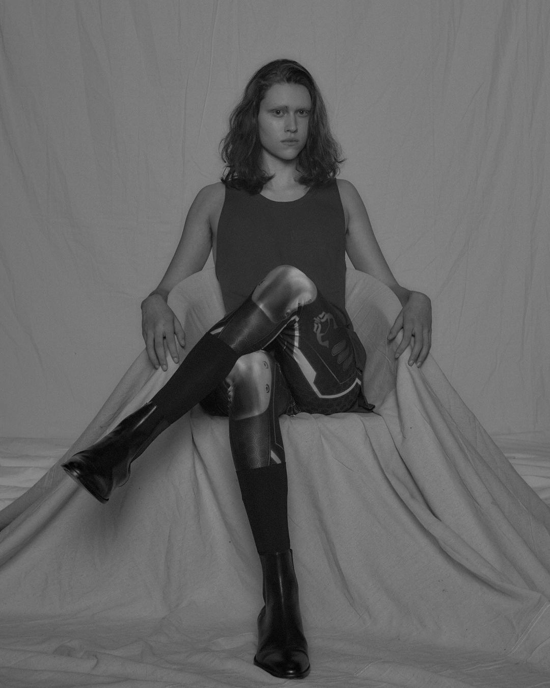 Monochromatic portrait of an androgynous person sitting in a draped chair wearing a tank top, printed tights, and black heeled boots.