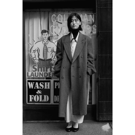 A young woman in a gray coat stands on a city sidewalk in front of a cleaners storefront, rendered in black and white.
