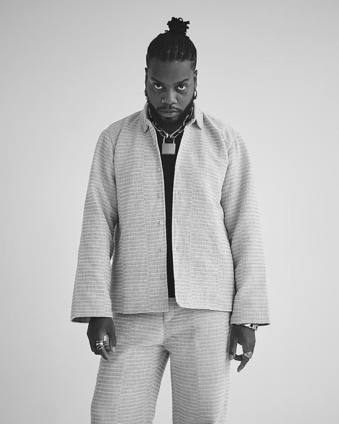 Black man in a textured off-white suit poses in a studio with a serious expression, wearing jewelry and a unique top knot hairstyle.