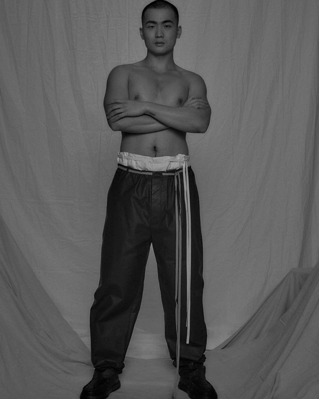 Monochromatic studio portrait of an adult Asian male posing with arms crossed, wearing dark pants, and a white fabric backdrop.