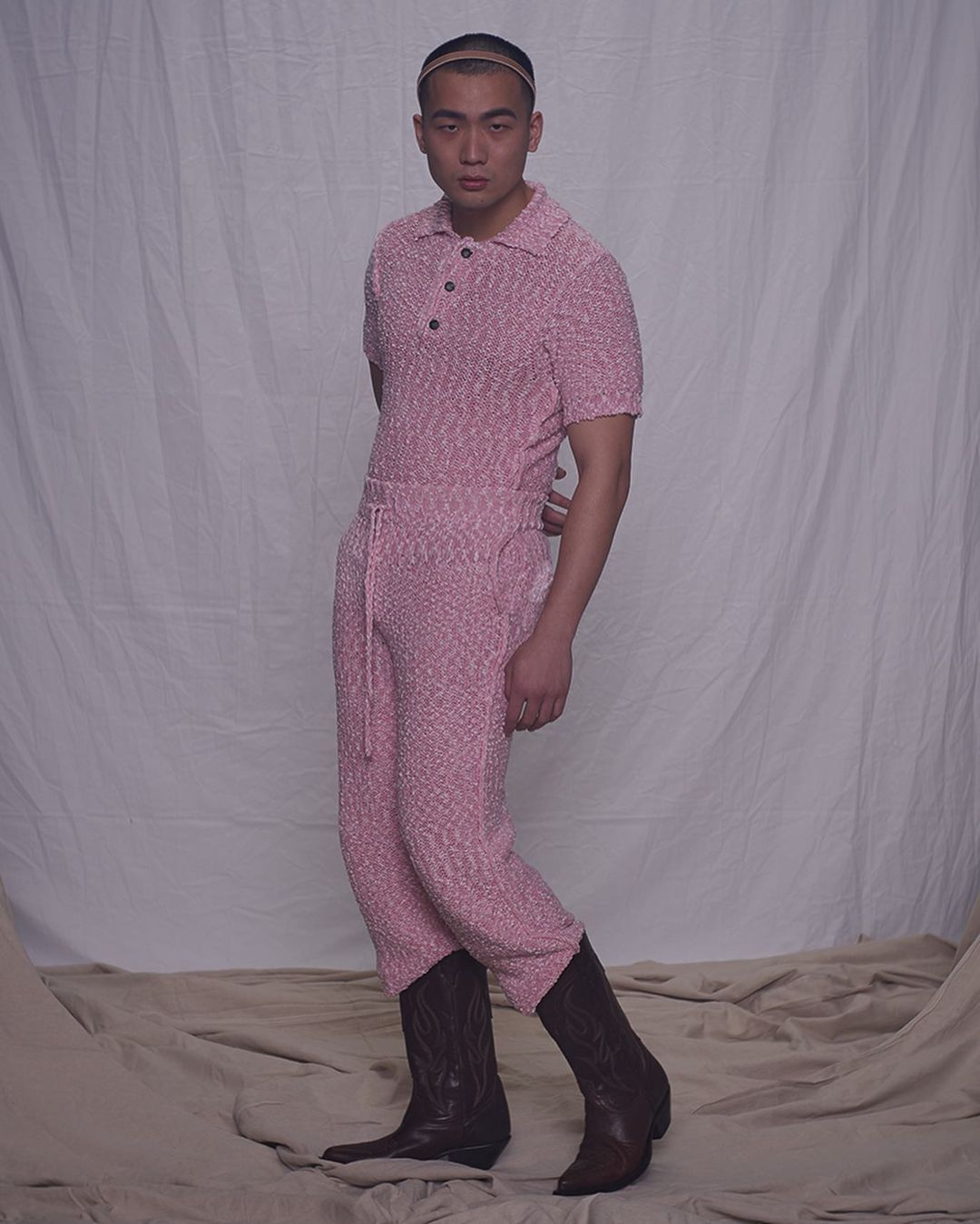Adult male poses indoors wearing pink knit outfit with brown cowboy boots, standing against a draped backdrop, medium shot.