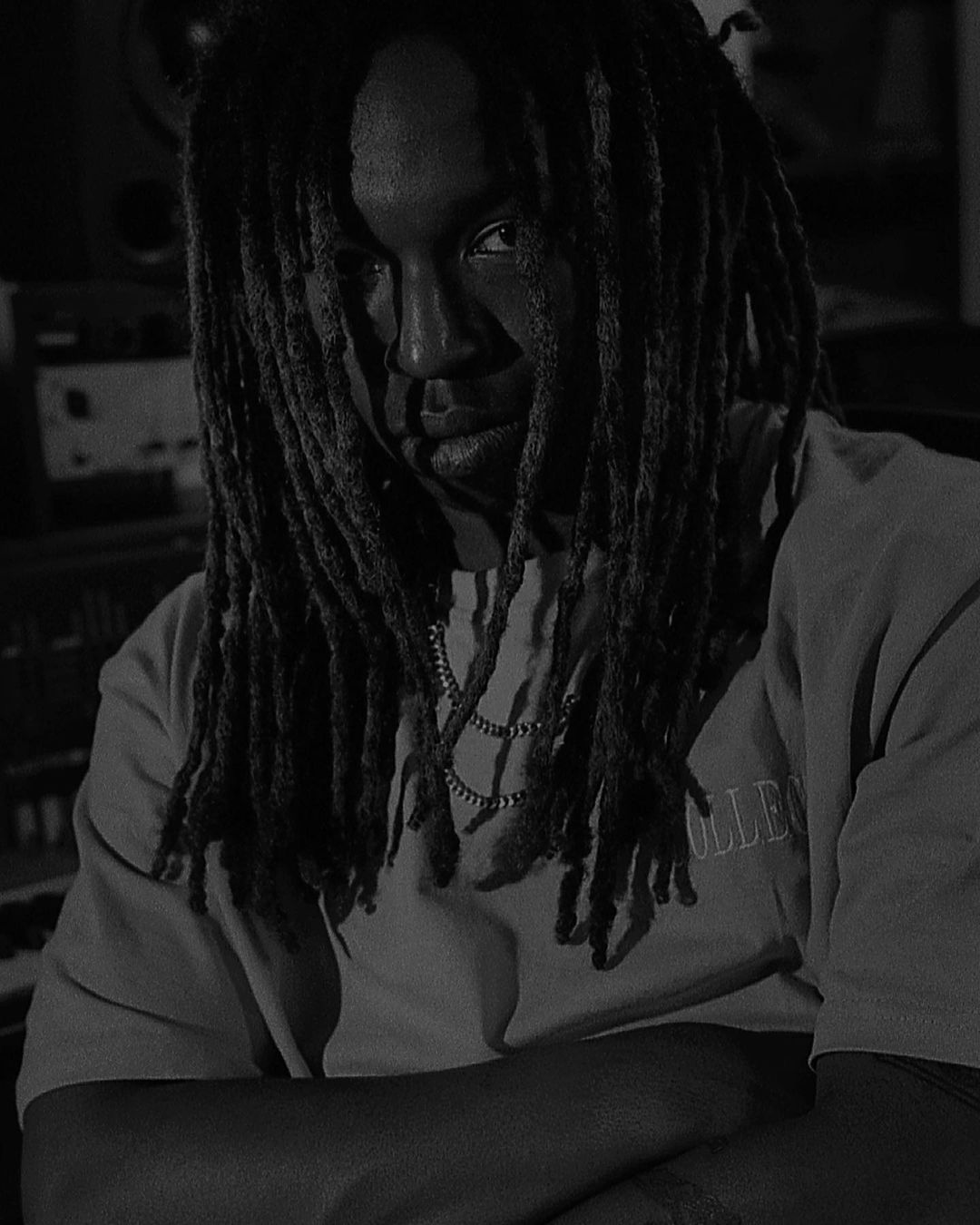 Monochrome portrait of a Black man with dreadlocks wearing a t-shirt and a chain, arms crossed, looking at the camera with a serious expression.