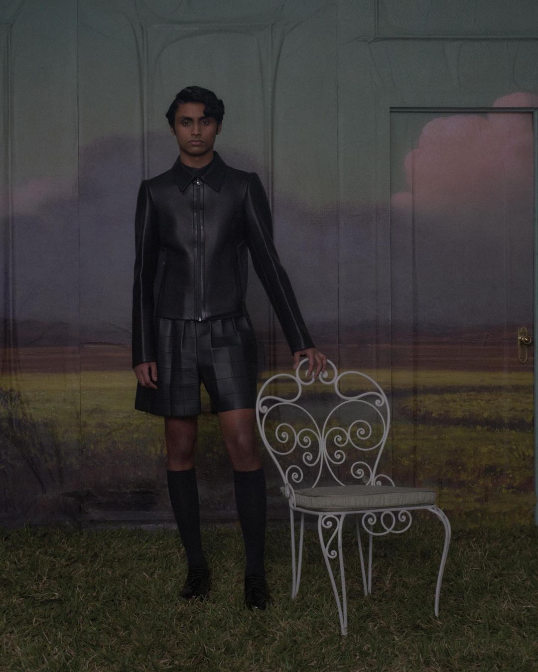 Adult male in black leather jacket and shorts stands next to a white decorative chair in front of a painted backdrop of clouds and grass.