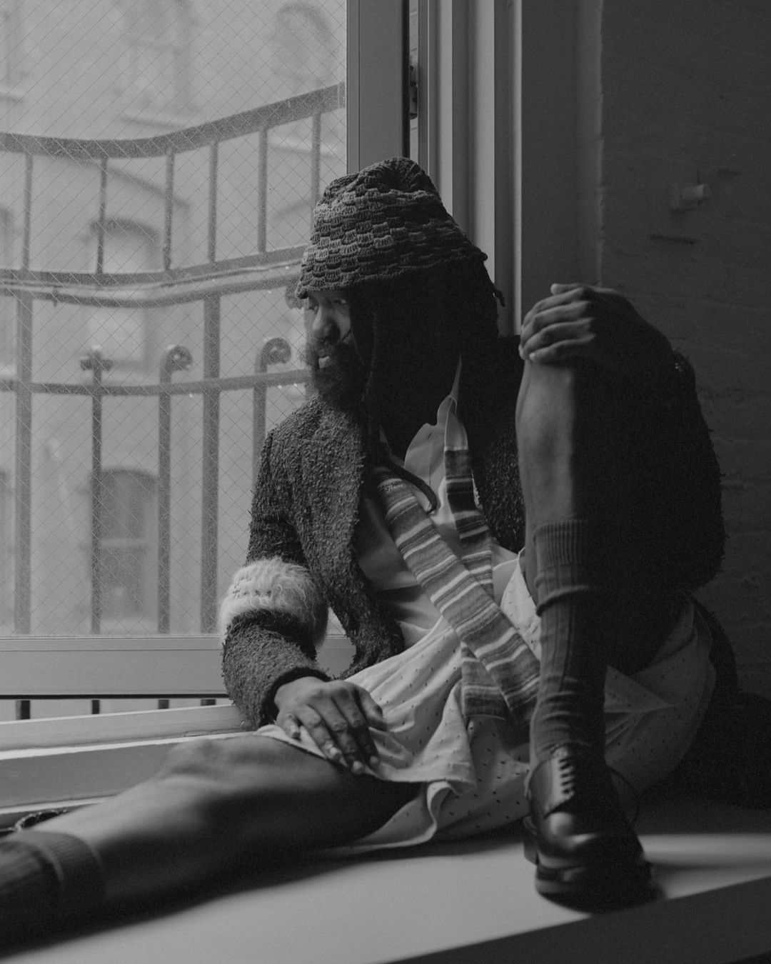 Black and white portrait of adult male in knit cap, sitting on windowsill with foot on wall, wearing jacket, dress, socks, and leather shoes.