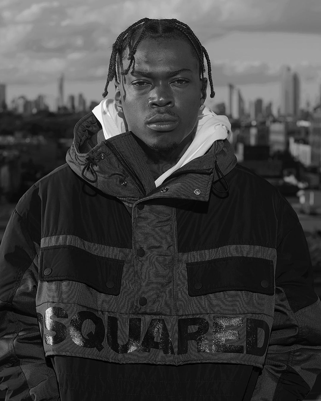 Black and white portrait of an African-American man with braided hair wearing a hooded jacket against an urban cityscape backdrop.