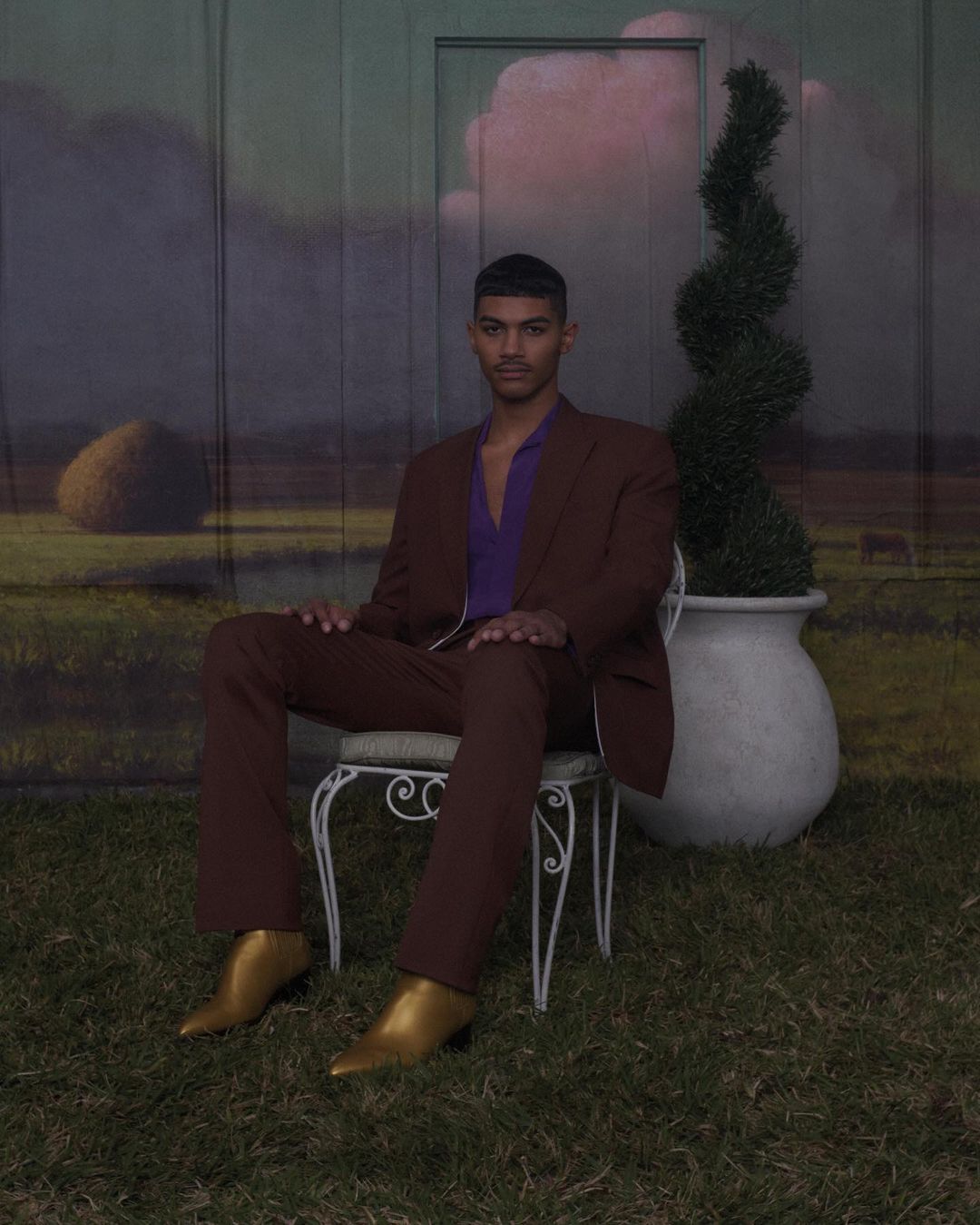 A man in a brown suit poses on a white chair against a painted outdoor backdrop, illuminated by soft lighting and styled with gold boots.