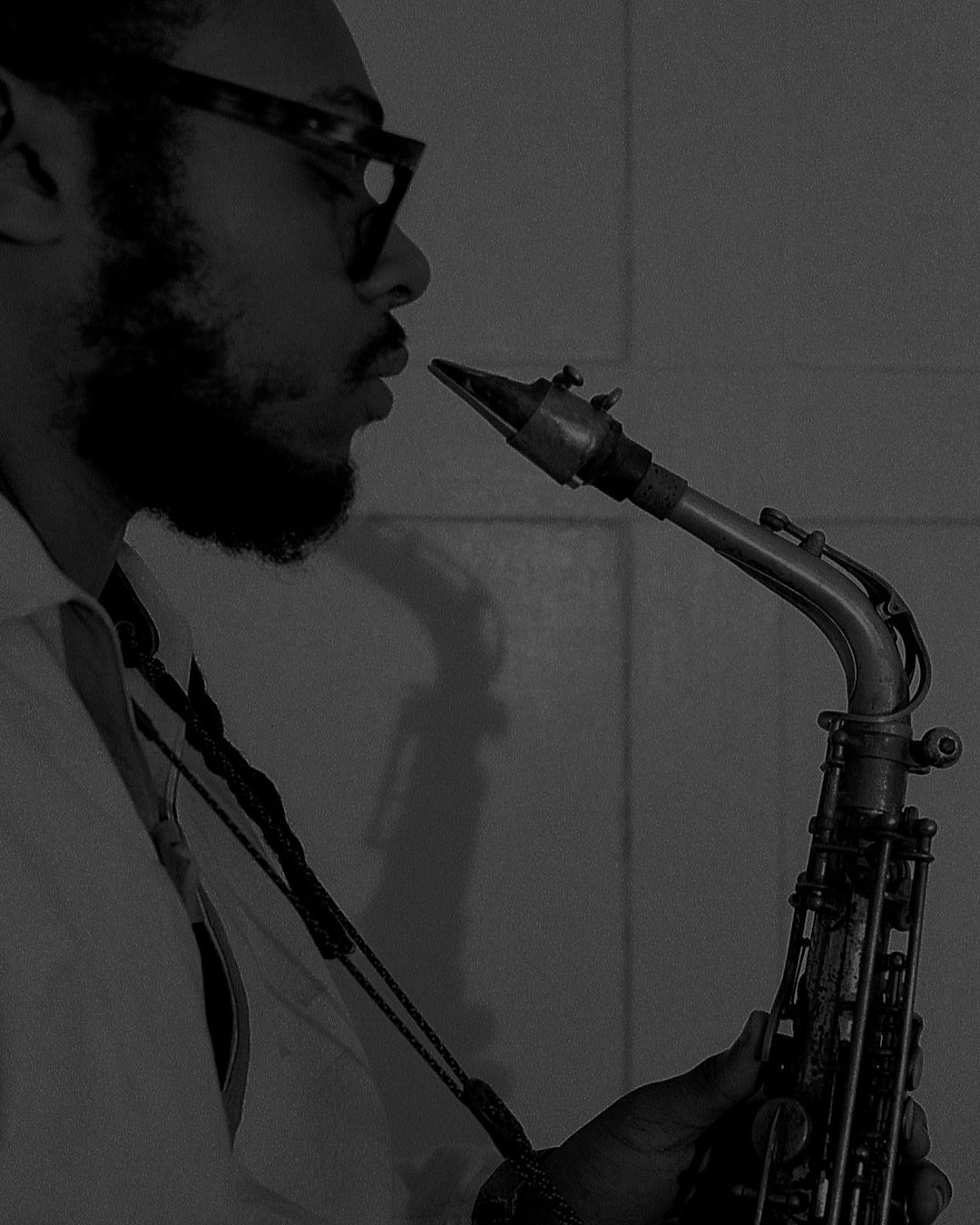 A man with glasses plays a saxophone, captured in a black and white studio shot with a close-up view emphasizing the instrument.