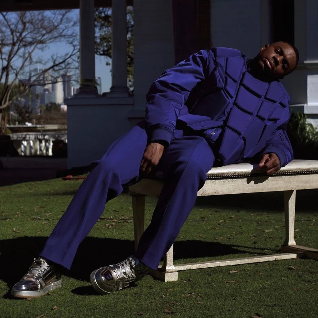 Portrait of an adult Black man wearing a blue jacket and pants, and metallic shoes. He is reclining on a bench outdoors.
