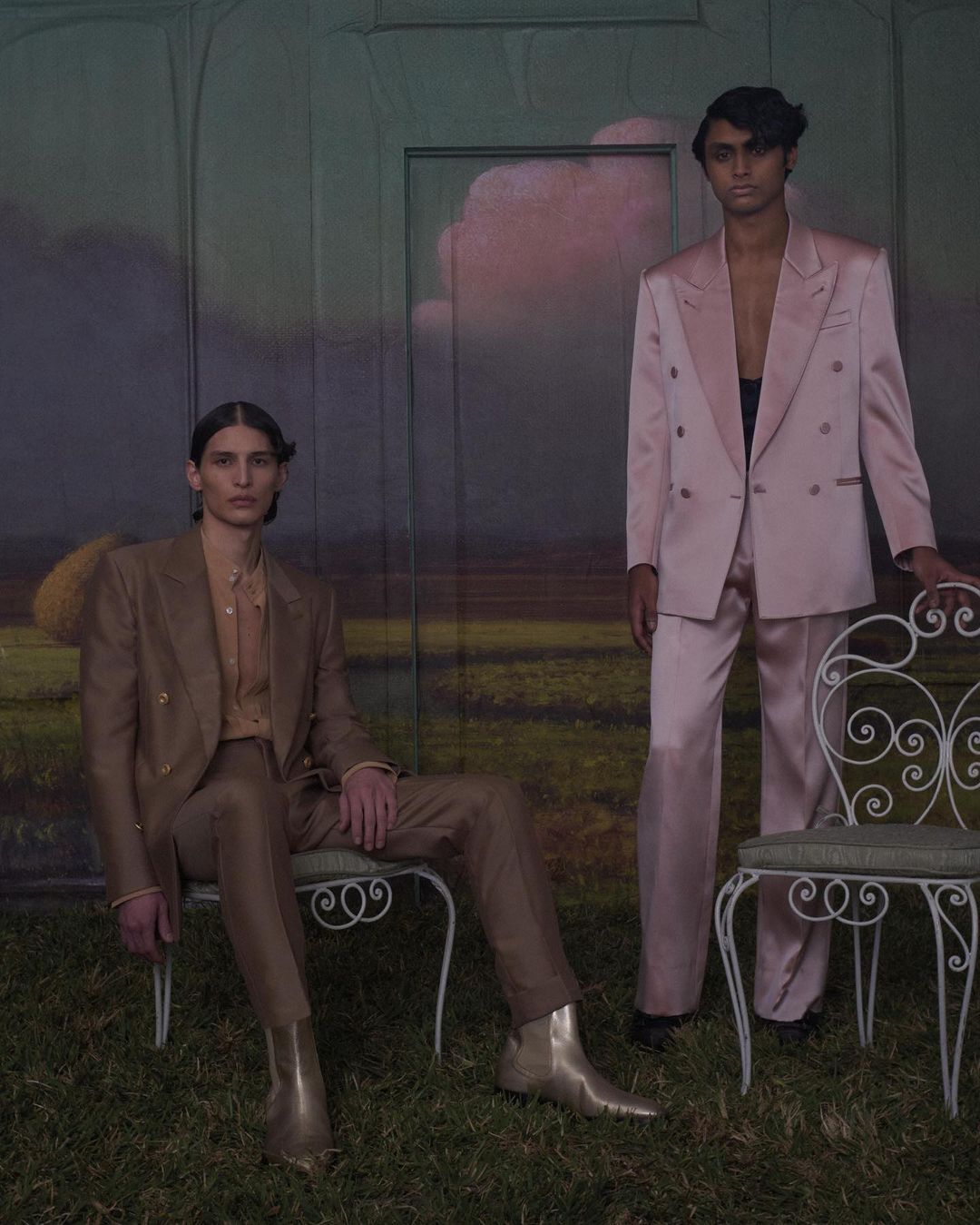 Two adults model tailored double-breasted suits in a photo studio with a surreal grassy set and cloud backdrop, seated and standing. 
