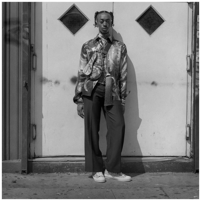 Black and white portrait of a slender man with braided hair, wearing a patterned shirt and black pants, posing in front of a textured white door.
