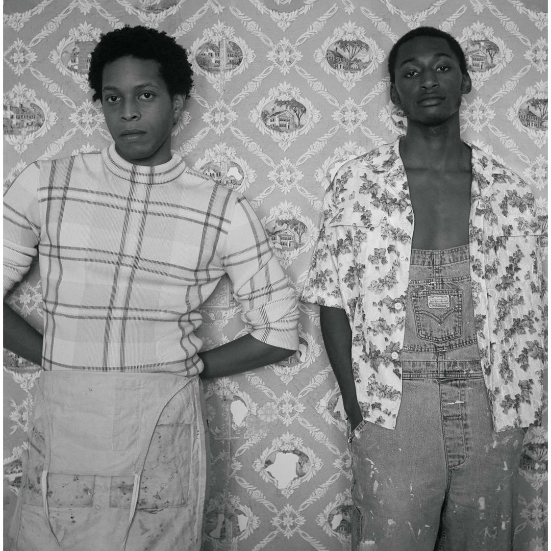 Black and white portrait of two young men in overalls, one in a plaid shirt, the other in a floral print shirt, posing against patterned wallpaper.