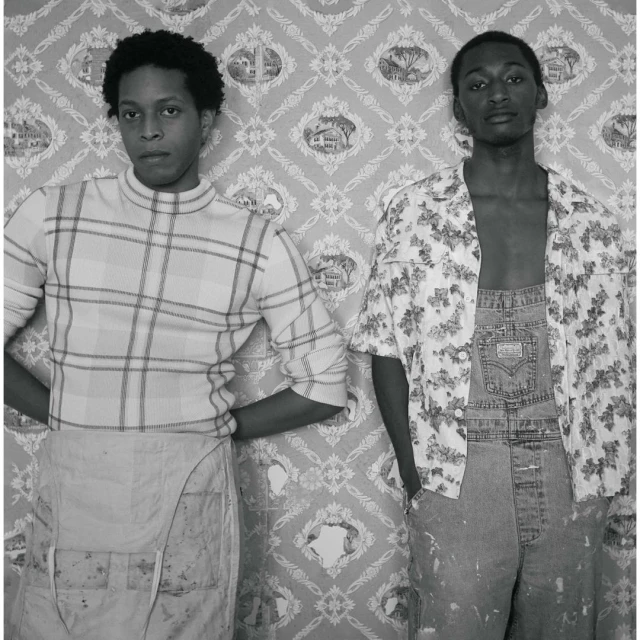 Black and white portrait of two young men in overalls, one in a plaid shirt, the other in a floral print shirt, posing against patterned wallpaper.