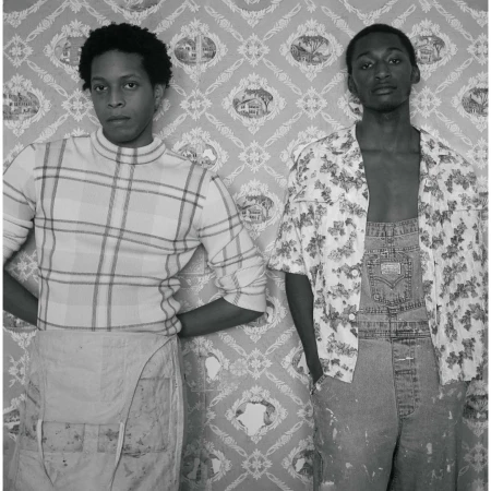 Black and white portrait of two young men in overalls, one in a plaid shirt, the other in a floral print shirt, posing against patterned wallpaper.