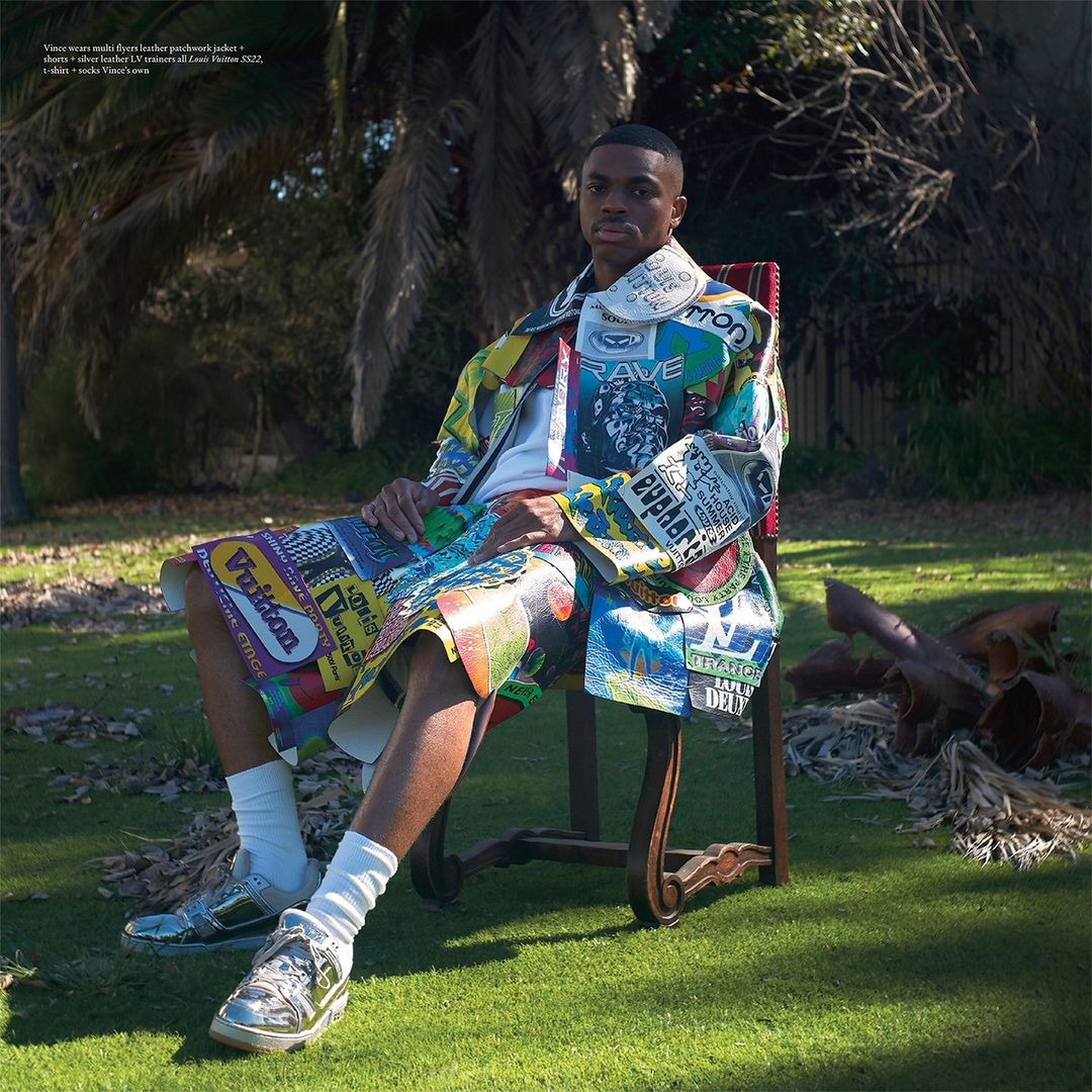 Man sits in a chair outdoors on grass, wearing a colorful patchwork jacket and shorts, white socks, and metallic sneakers in a daytime setting.