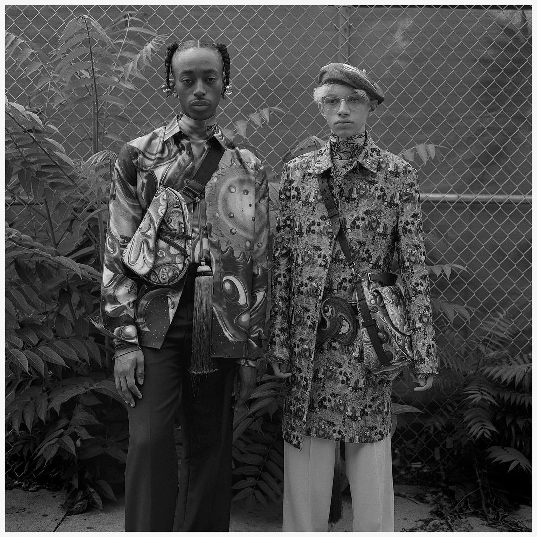 Two fashion models pose outdoors in patterned shirts, wide-leg pants, and shoulder bags against a chain-link fence and leafy green plants.