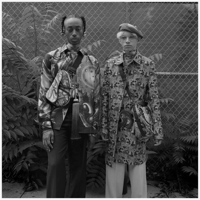 Two fashion models pose outdoors in patterned shirts, wide-leg pants, and shoulder bags against a chain-link fence and leafy green plants.