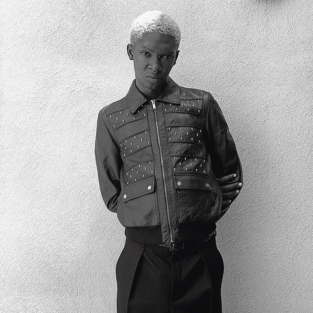 Black and white portrait of an adult with short, bleached blonde hair wearing a jacket with detailing against a textured wall.