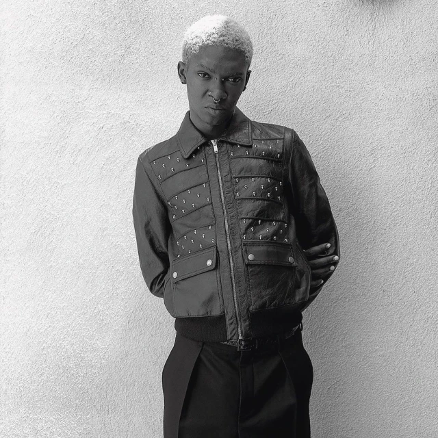 Black and white portrait of an adult with short, bleached blonde hair wearing a jacket with detailing against a textured wall.