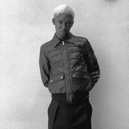 Black and white portrait of an adult with short, bleached blonde hair wearing a jacket with detailing against a textured wall.