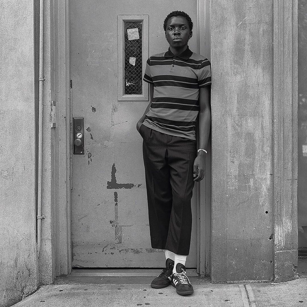 A young Black man leans against a weathered doorway in a medium shot, showcasing a striped polo shirt, dark trousers, and sneakers.