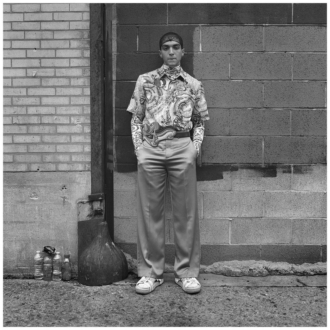 A man in patterned shirt and light pants stands against a brick wall. He has his hands in his pockets in this full outdoor shot.