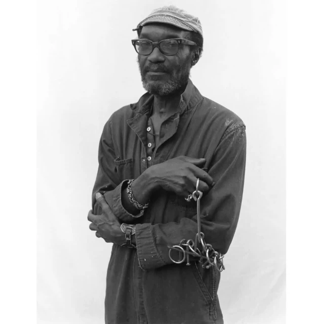 Black and white portrait of an adult man wearing glasses and a cap, with metal rings attached to his wrist, against a neutral background.