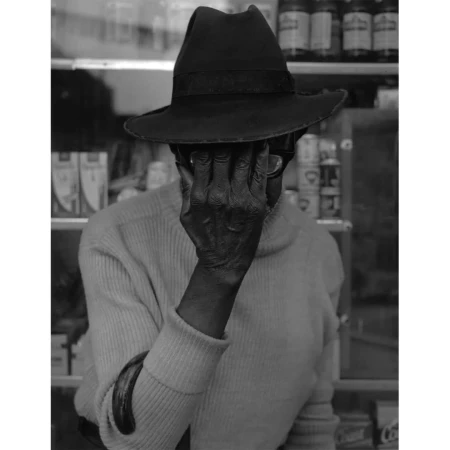 Black and white portrait of a person wearing a hat and glasses, obscuring their face with their hand in an indoor setting.