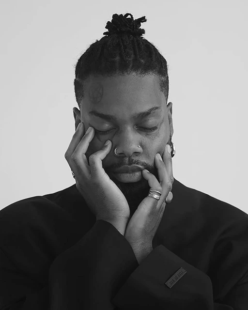 Black and white close-up portrait of a Black adult male with dreadlocks touching his face, wearing a dark jacket, against a white background.