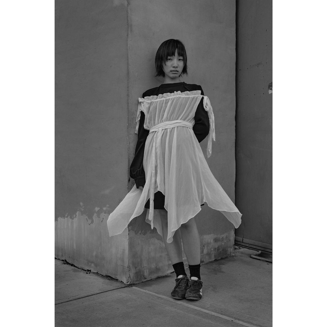 Monochrome fashion portrait of an Asian young adult woman in a white dress posing against a concrete wall, showcasing unique style.
