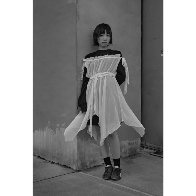 Monochrome fashion portrait of an Asian young adult woman in a white dress posing against a concrete wall, showcasing unique style.