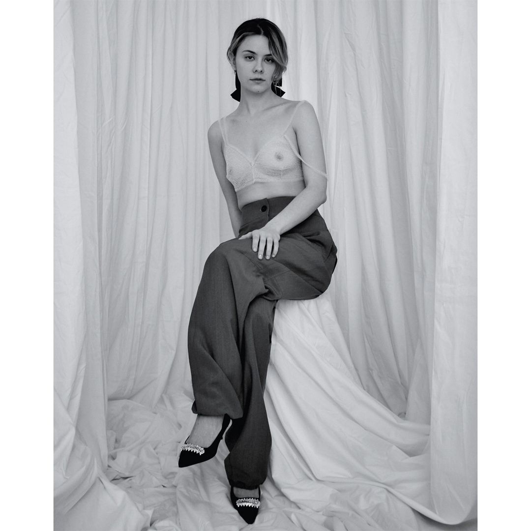 Black and white portrait of a young woman with a serious facial expression, wearing a bra top and wide leg pants, posing against a white backdrop.