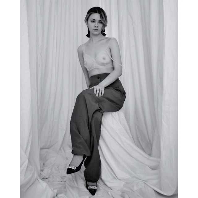 Black and white portrait of a young woman with a serious facial expression, wearing a bra top and wide leg pants, posing against a white backdrop.