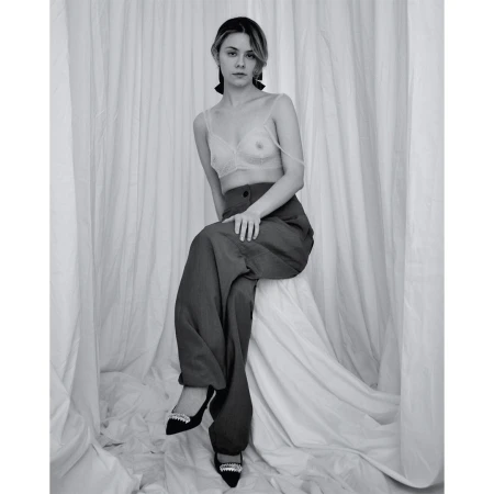 Black and white portrait of a young woman with a serious facial expression, wearing a bra top and wide leg pants, posing against a white backdrop.