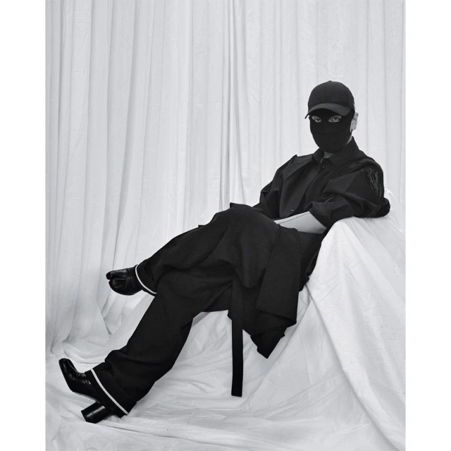 Person wearing a black balaclava and baseball cap seated in a chair with a draped cloth, wearing black clothing and boots in photo studio.
