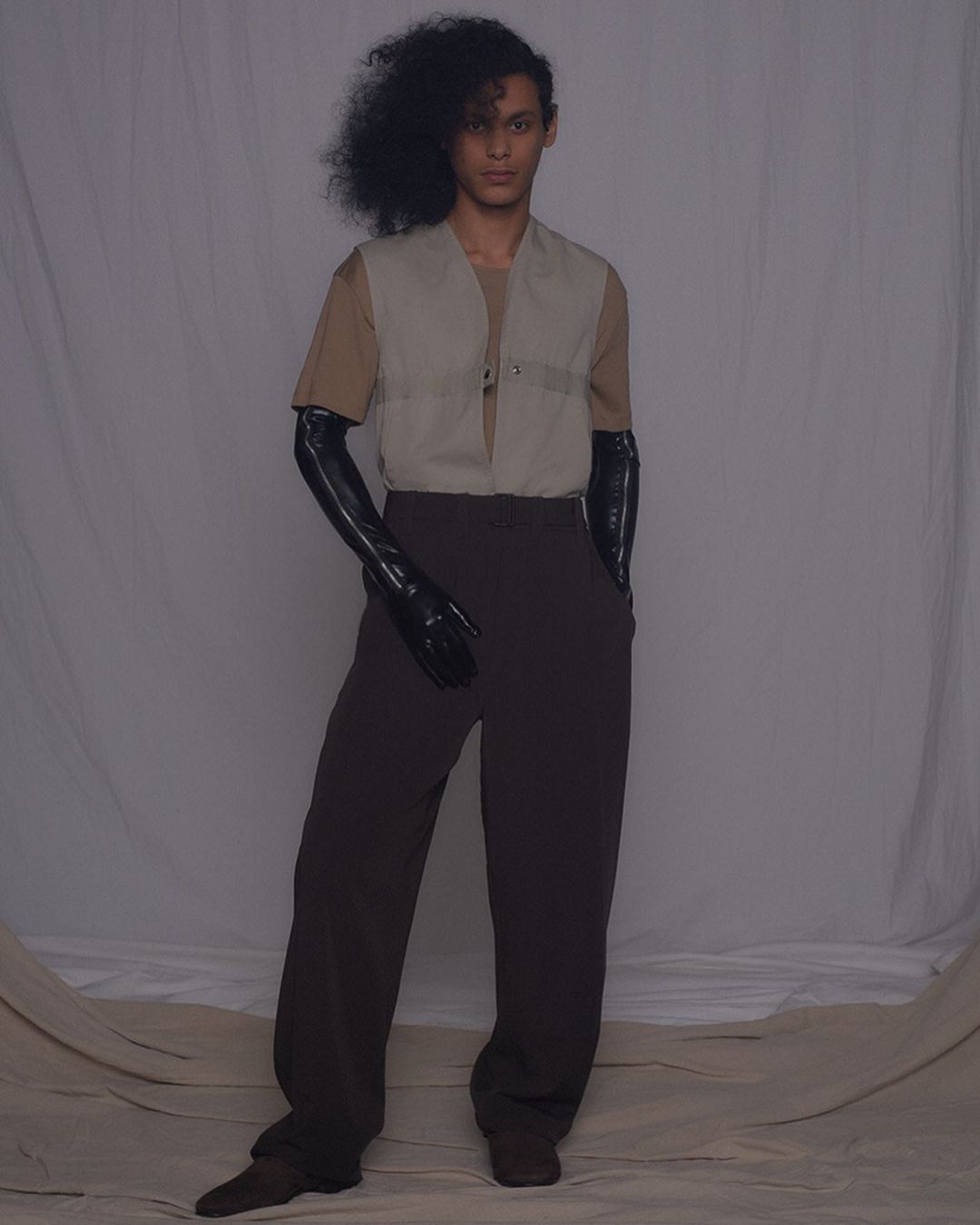 Fashionable young adult poses in an earth-toned T-shirt, beige vest, dark pants, and high black gloves against a simple studio backdrop.