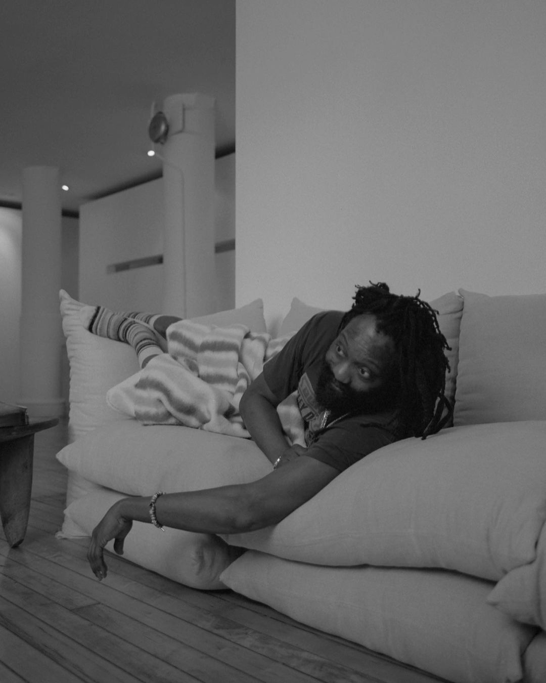 Monochrome portrait of a Black adult male with dreadlocks, lying casually on a white couch, looking at the camera with a calm facial expression.