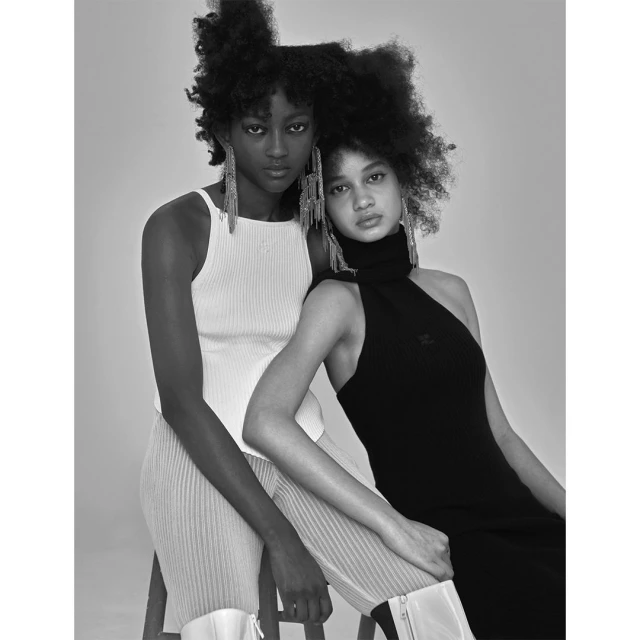 Monochromatic portrait of two women with afro hairstyles wearing dresses and large metallic earrings, posing in a photo studio.