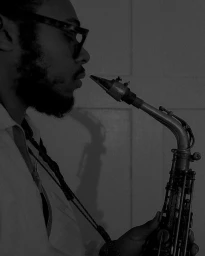 Black man wearing glasses and a light shirt playing a saxophone in profile view.