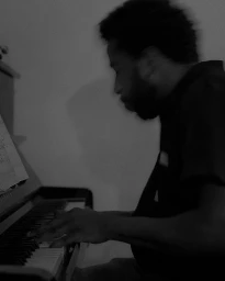 Black and white profile portrait of a black man playing the piano in a dimly lit indoor space, captured in London by Katsu Naito.