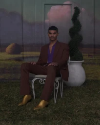 Man in a brown suit and purple shirt seated on a white wire chair wearing metallic gold boots against a painted landscape backdrop.