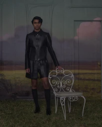 A man in a black leather jacket and shorts stands next to an ornate white metal chair against a painted landscape backdrop.