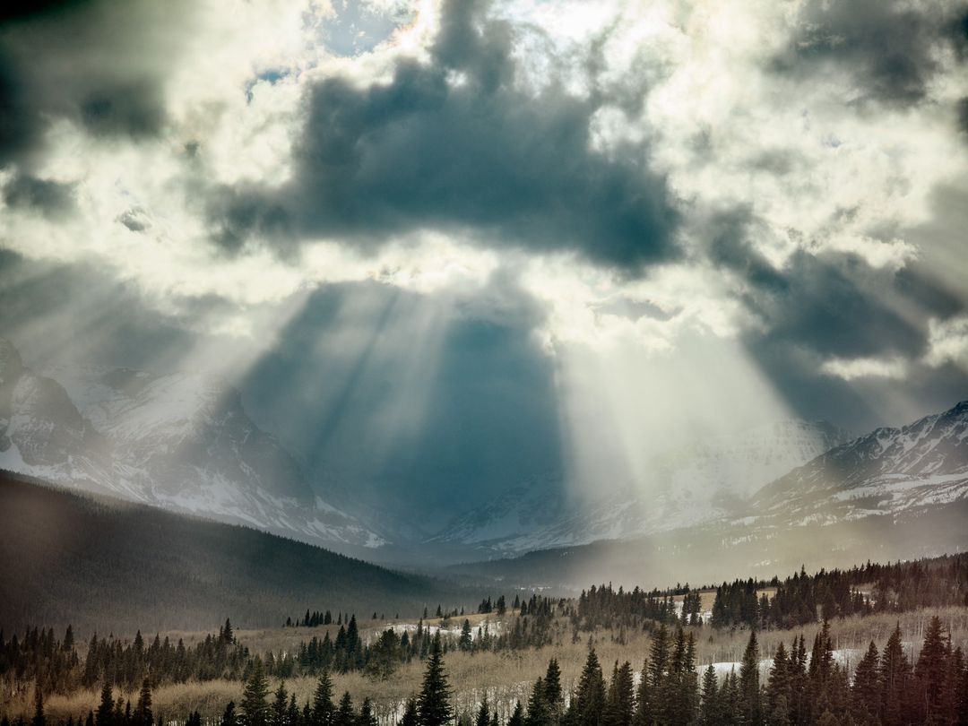 Sun rays streaming through dark clouds over a mountain range with pine trees. Moody nature photograph of light and shadow, forest landscape.