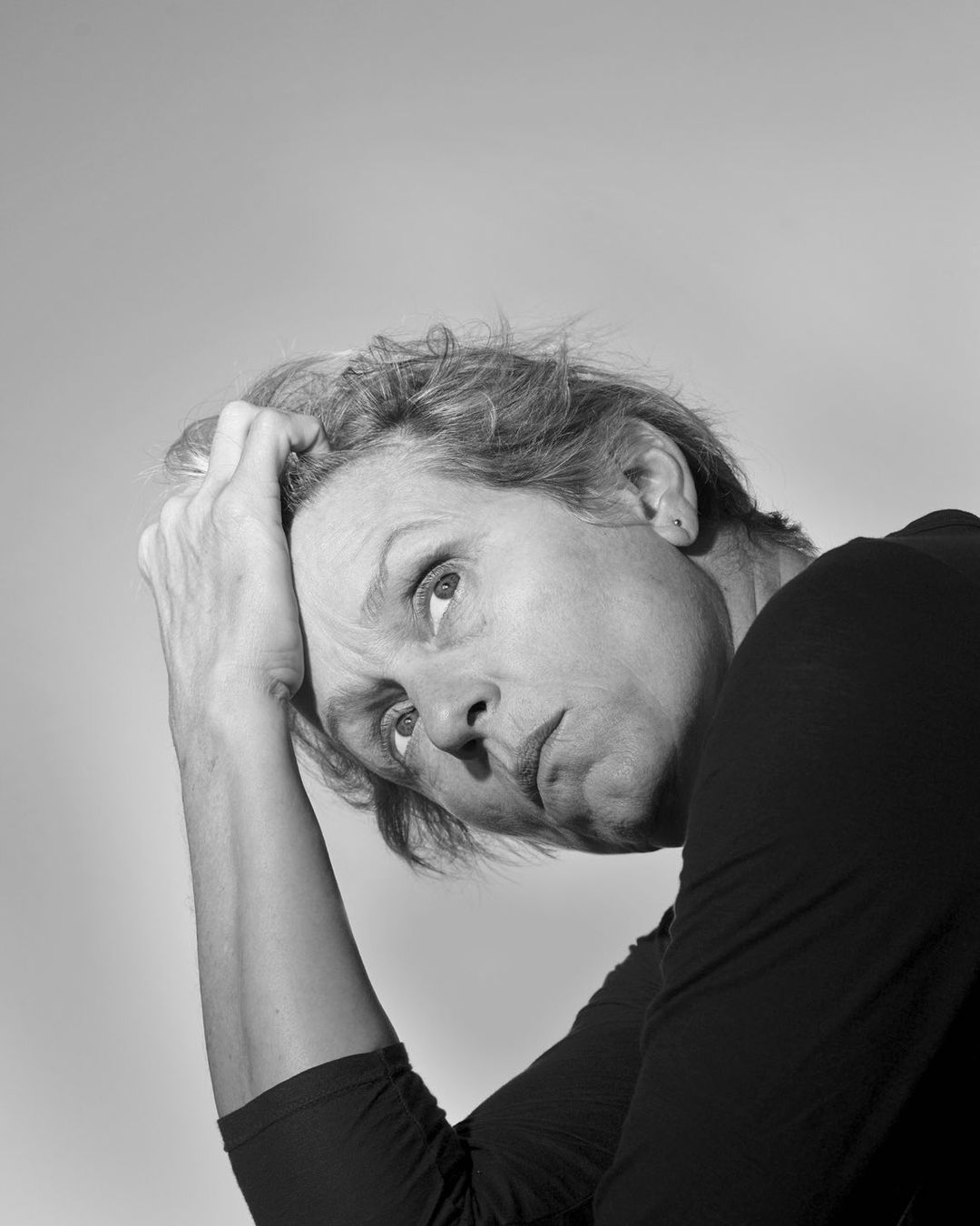 Black and white portrait of a mature woman with short hair resting her head in her hand, looking upwards in a simple, neutral background.