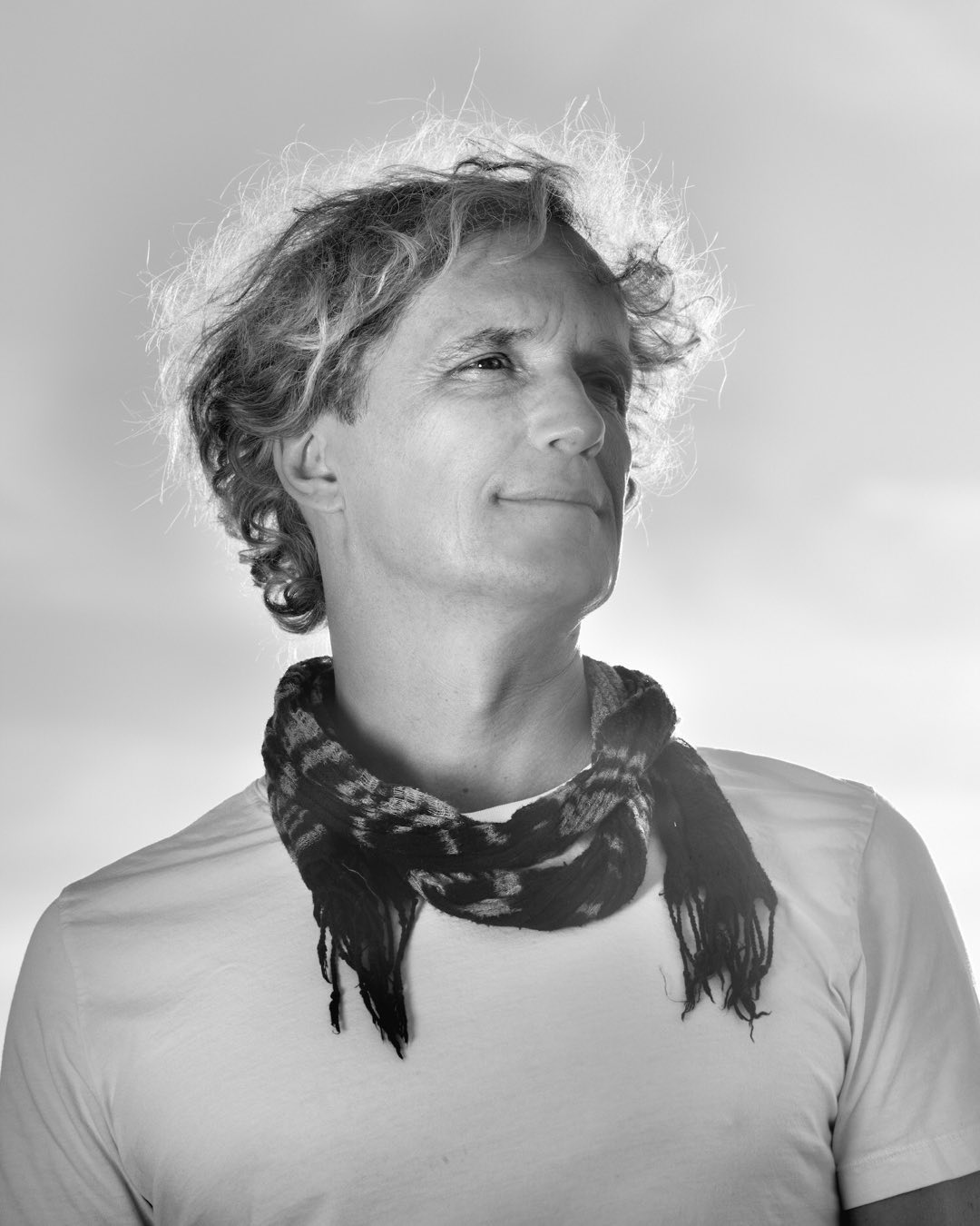 Black and white portrait of an adult man wearing a t-shirt and patterned scarf, looking off camera, against a bright, light background.