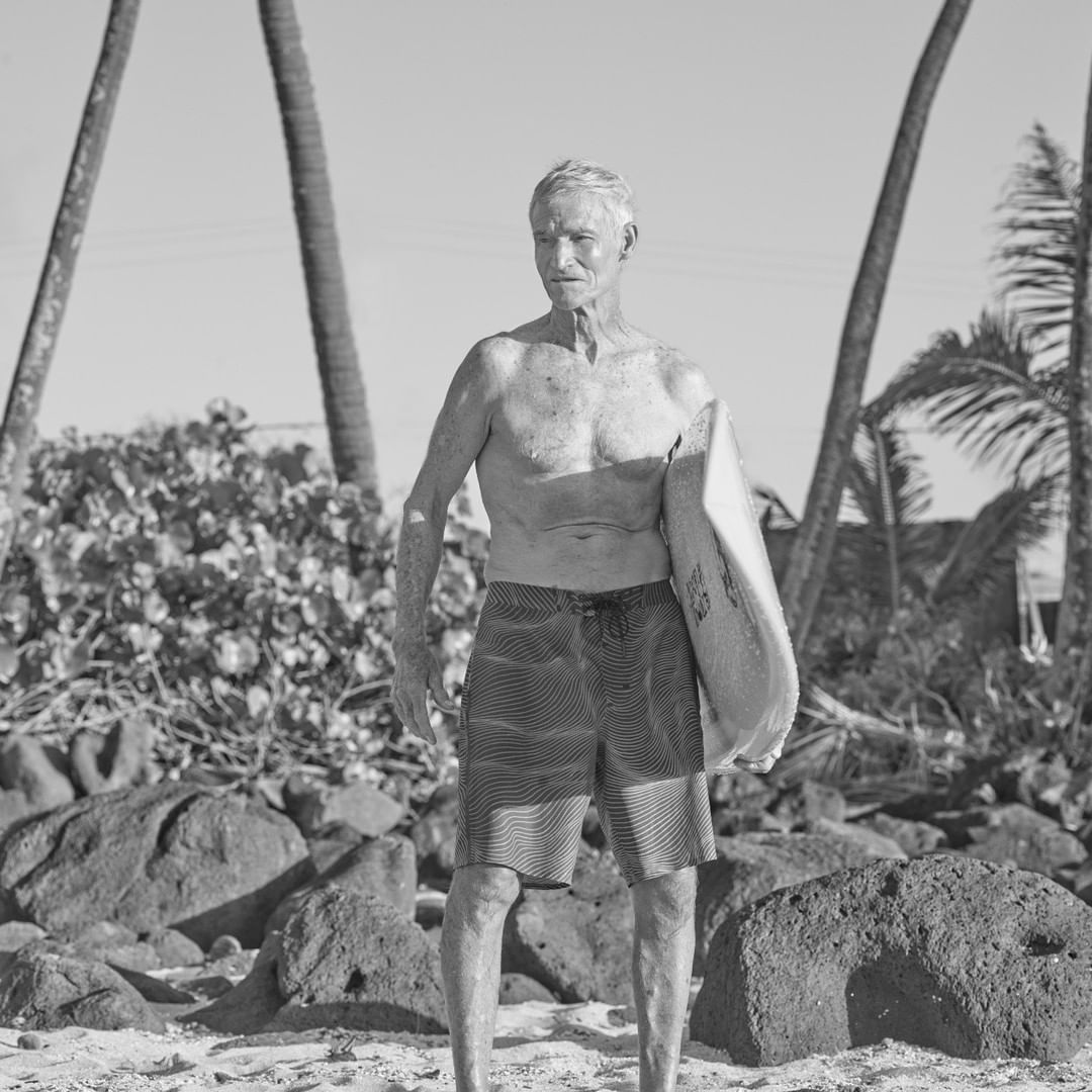 Older man on a beach in black and white, shirtless, holding a surfboard with palm trees and vegetation in the background.