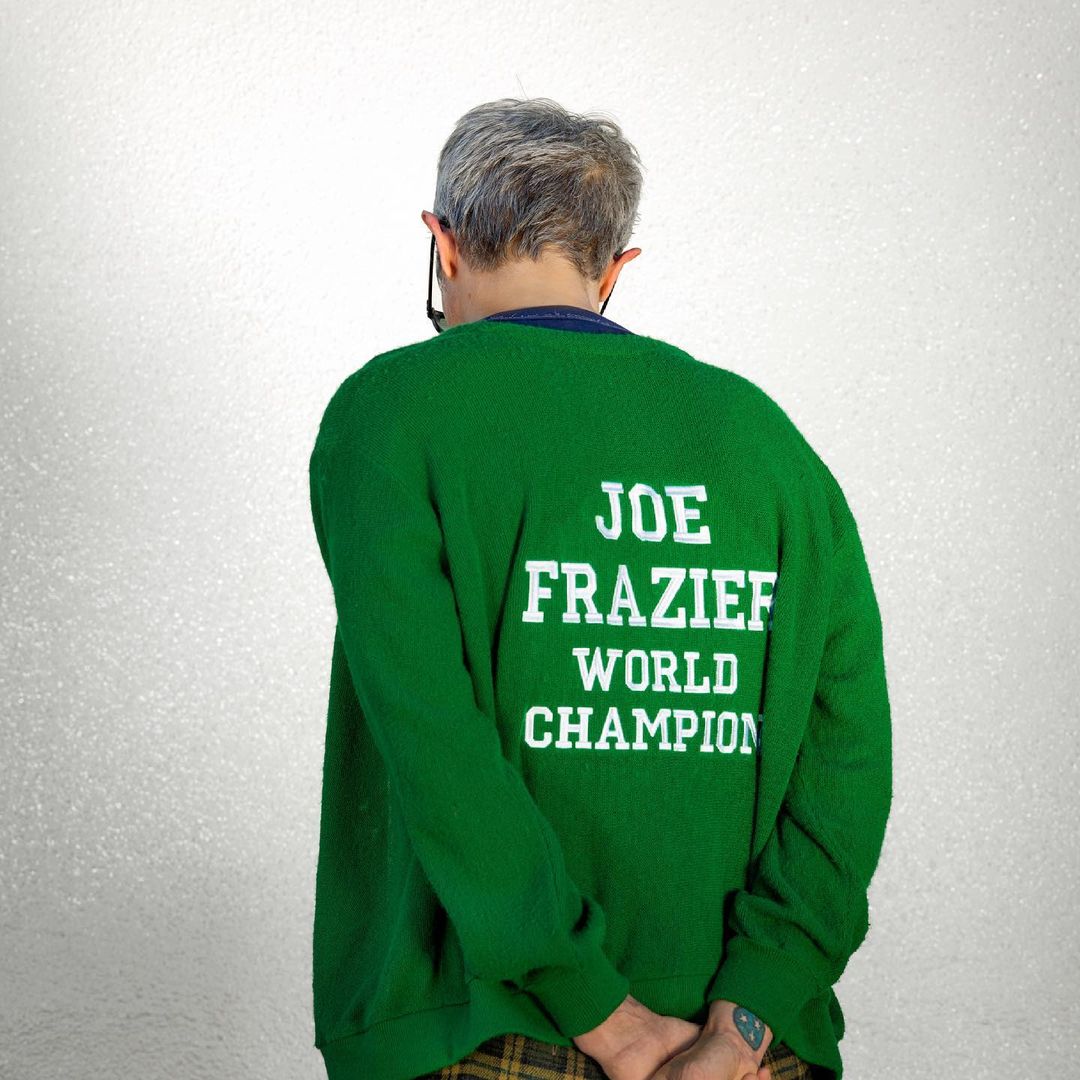 An adult with gray hair is shown from the rear wearing a green sweater with the words 'Joe Frazier World Champion' in white text.