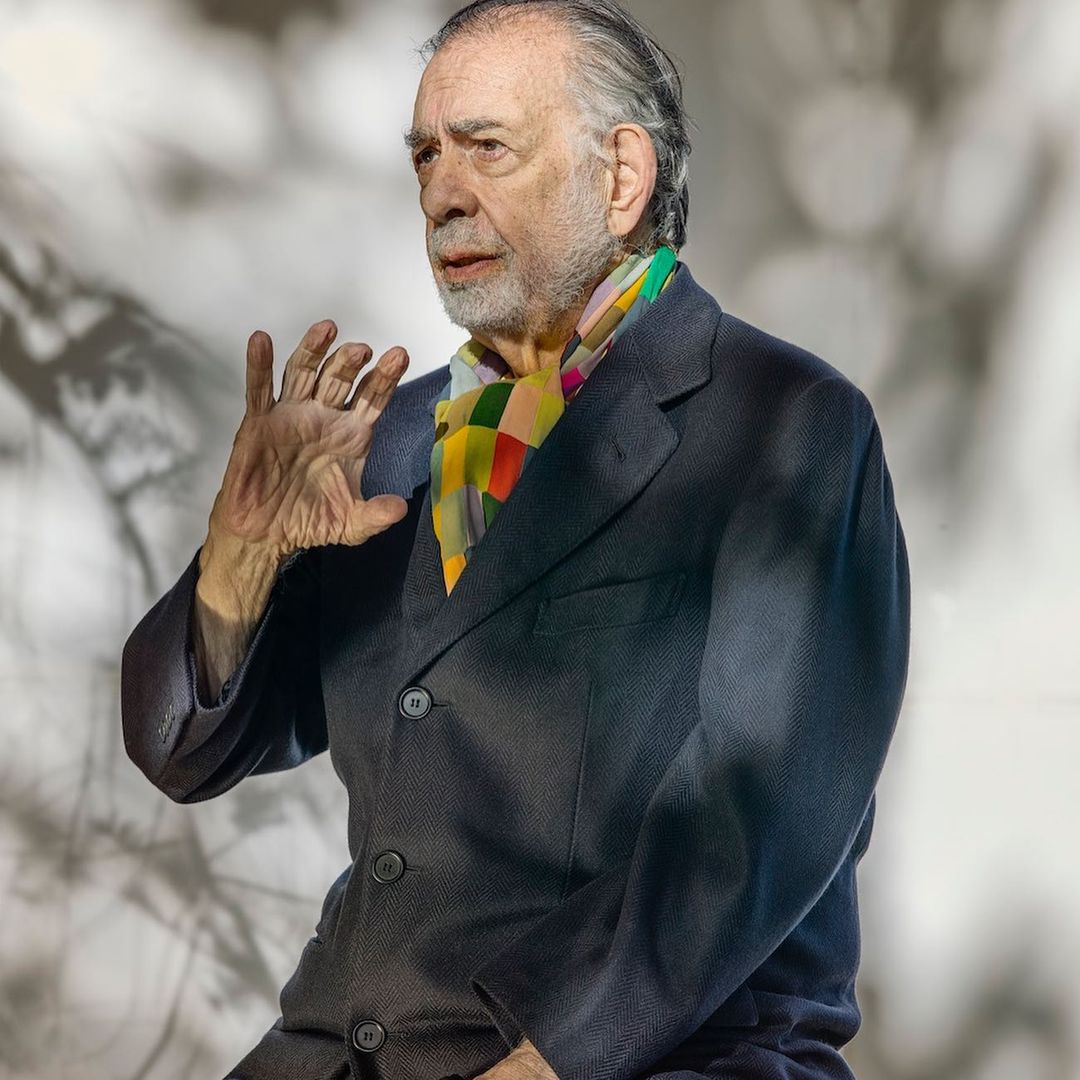 Older adult male wearing a blazer and patterned scarf, seated indoors with a blurred light background and gesturing with his left hand.